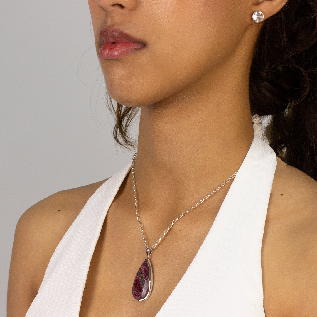 Woman wearing a silver teardrop-shaped pinky-red crystal pendant on a silver chain against a plain background