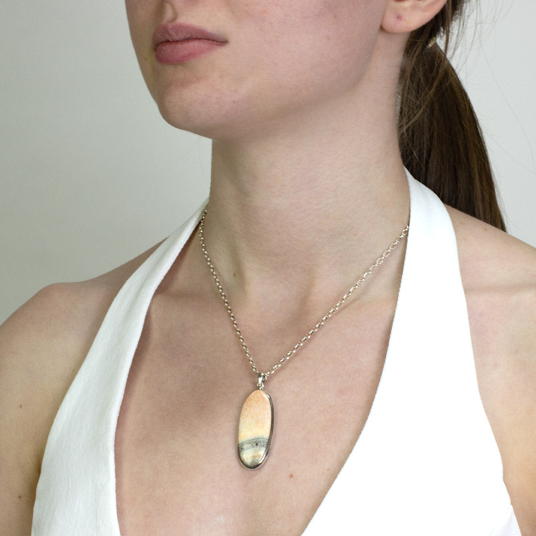 Woman wearing a silver necklace with a stone with salmon-pink tones and soft grey banding on a plain background