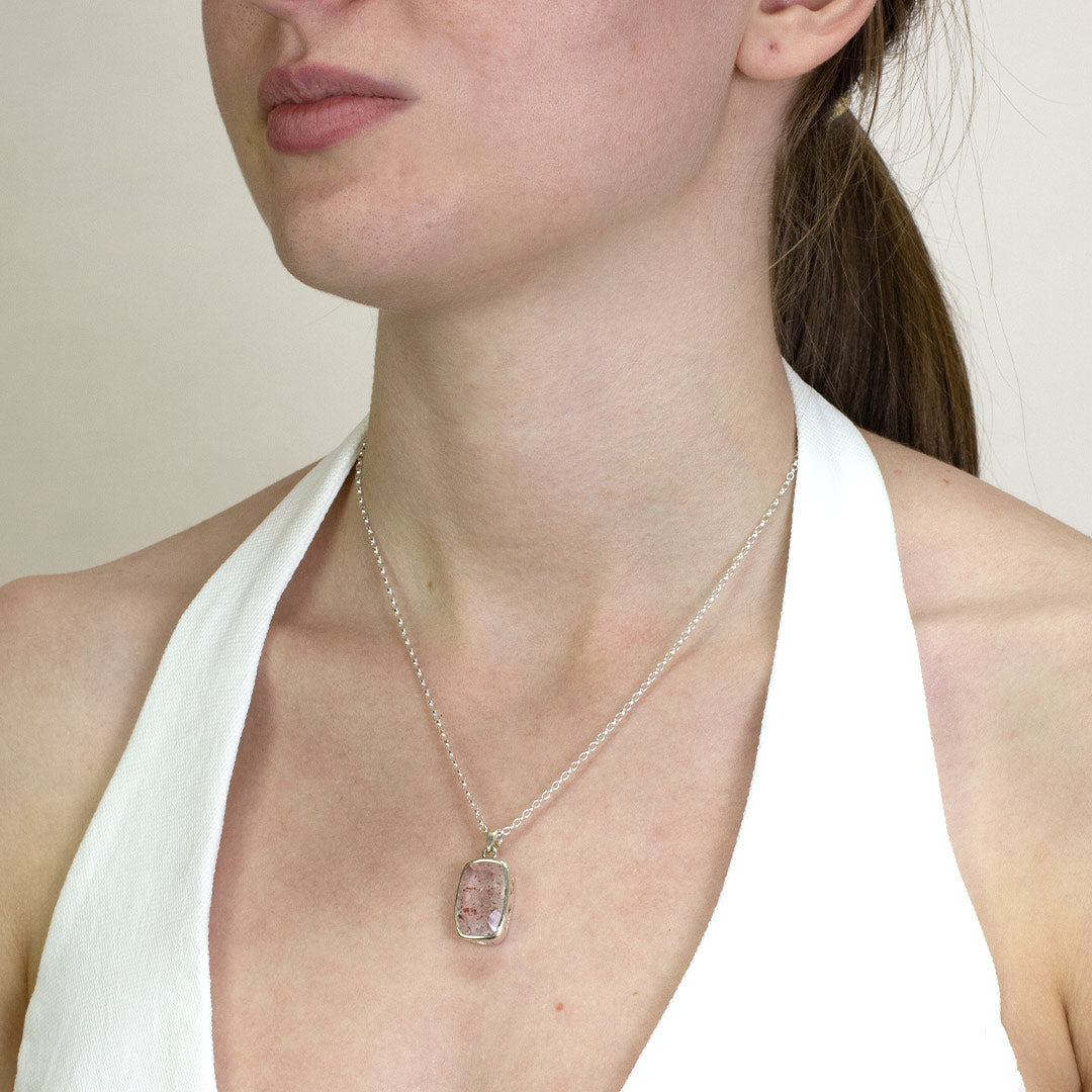 Woman wearing a silver necklace with a clear stone with pink and red inclusions on a plain background