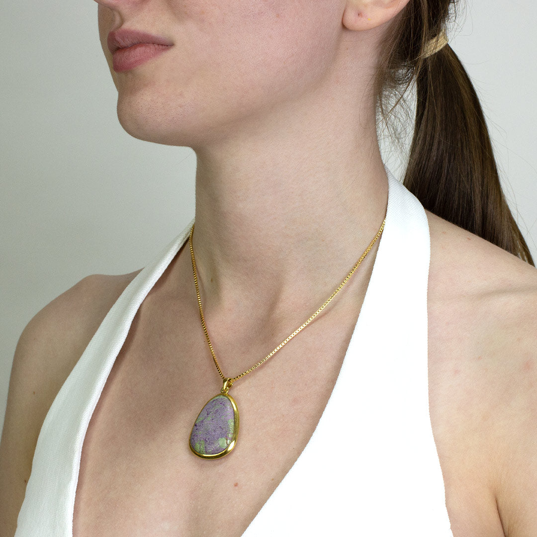 Woman wearing a gold necklace with a purple and green stone on a plain background