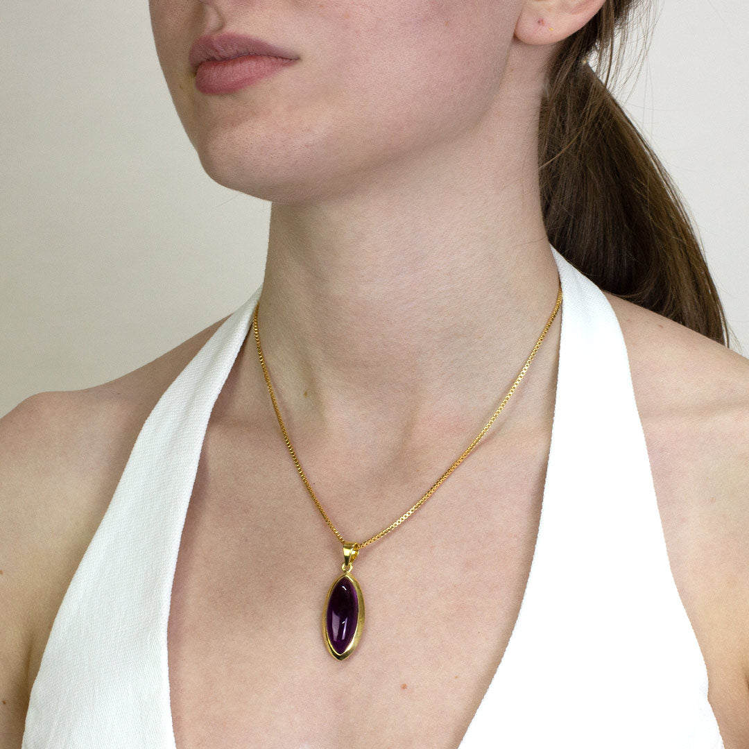 Woman wearing a gold necklace with a purple gemstone on a plain background