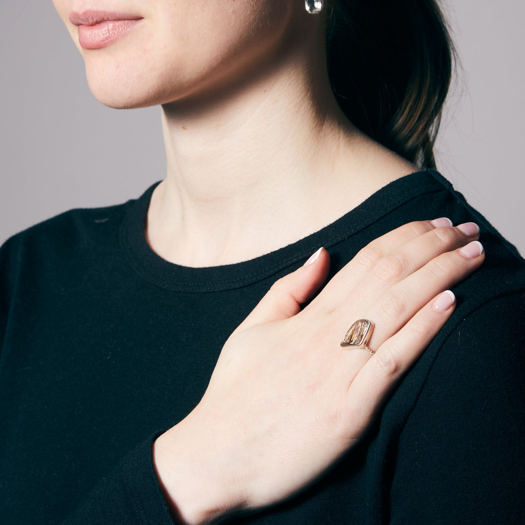 Close-up of a person wearing a black shirt with a hand resting on their shoulder, showing a silver ring with a metallic stone on a plain background.