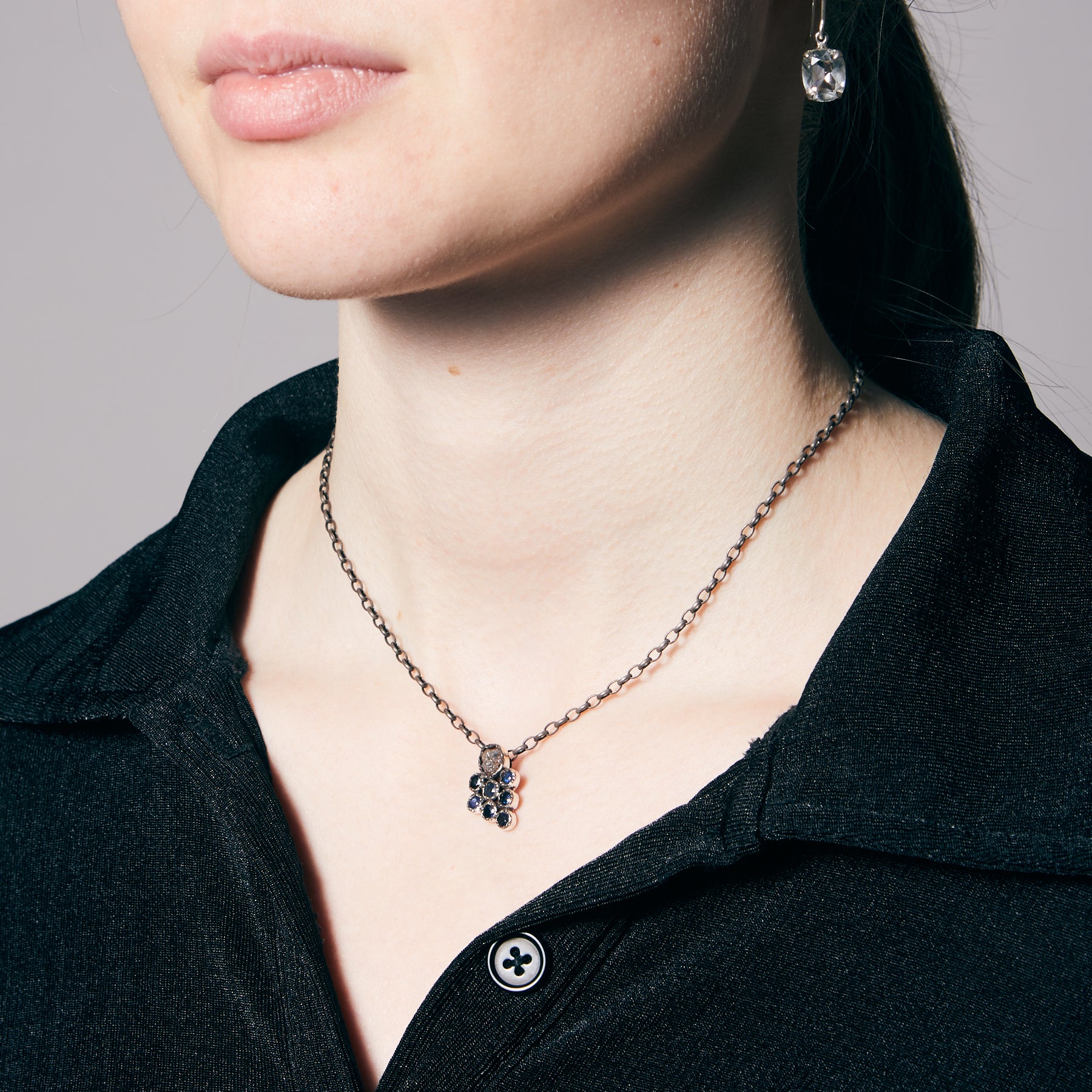 Person wearing a necklace with a grape shaped pendant featuring blue gemstones on a neutral background