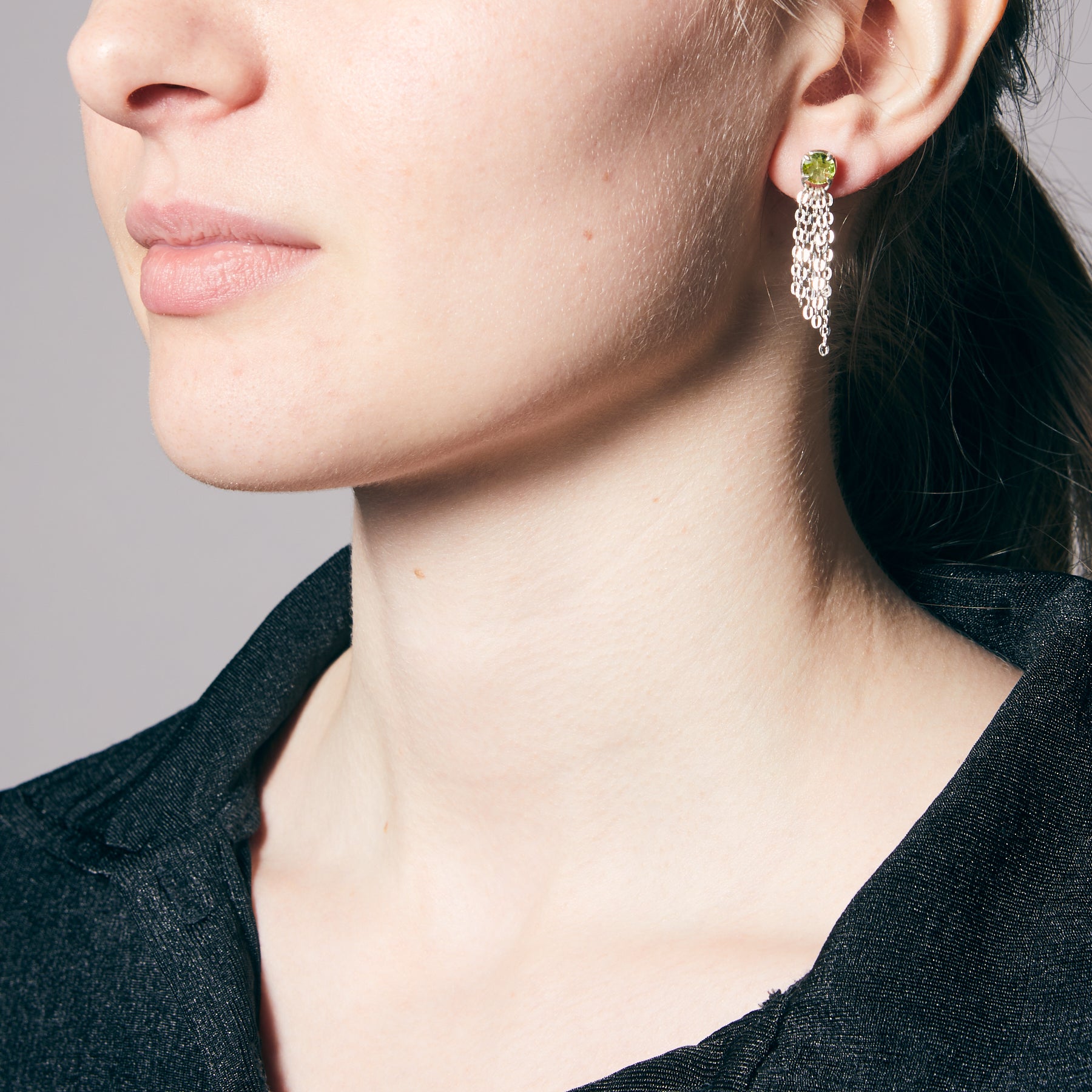 Close-up of a person wearing a silver chain earring with a green gemstone on a neutral background