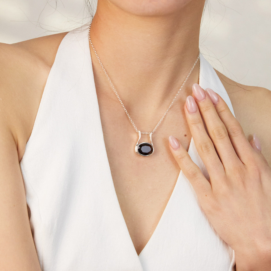 A close-up of a model wearing a silver necklace featuring a pendant with a brown gemstone, against a plain background