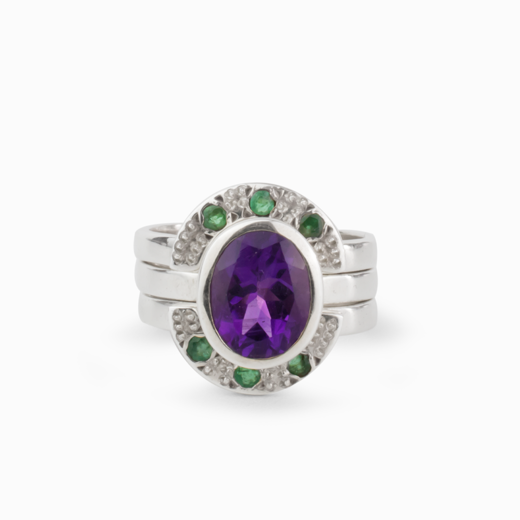3 ring bridal band, middle ring is large faceted bezel set amethyst. The rings above and below both curve around the amethyst containing 3 smaller faceted bezel set emeralds. All coming together to make one ring.