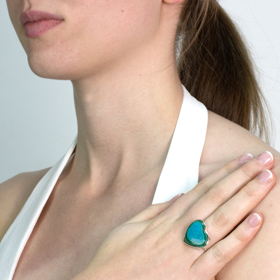 Woman wearing a silver ring with a heart-shaped green and blue gemstone on a plain background