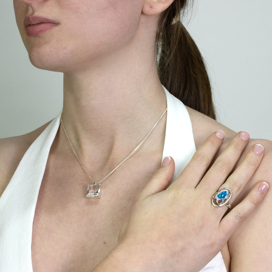 Woman wearing a silver ring with an oval stone featuring a blue centre and brown edges on a plain background.