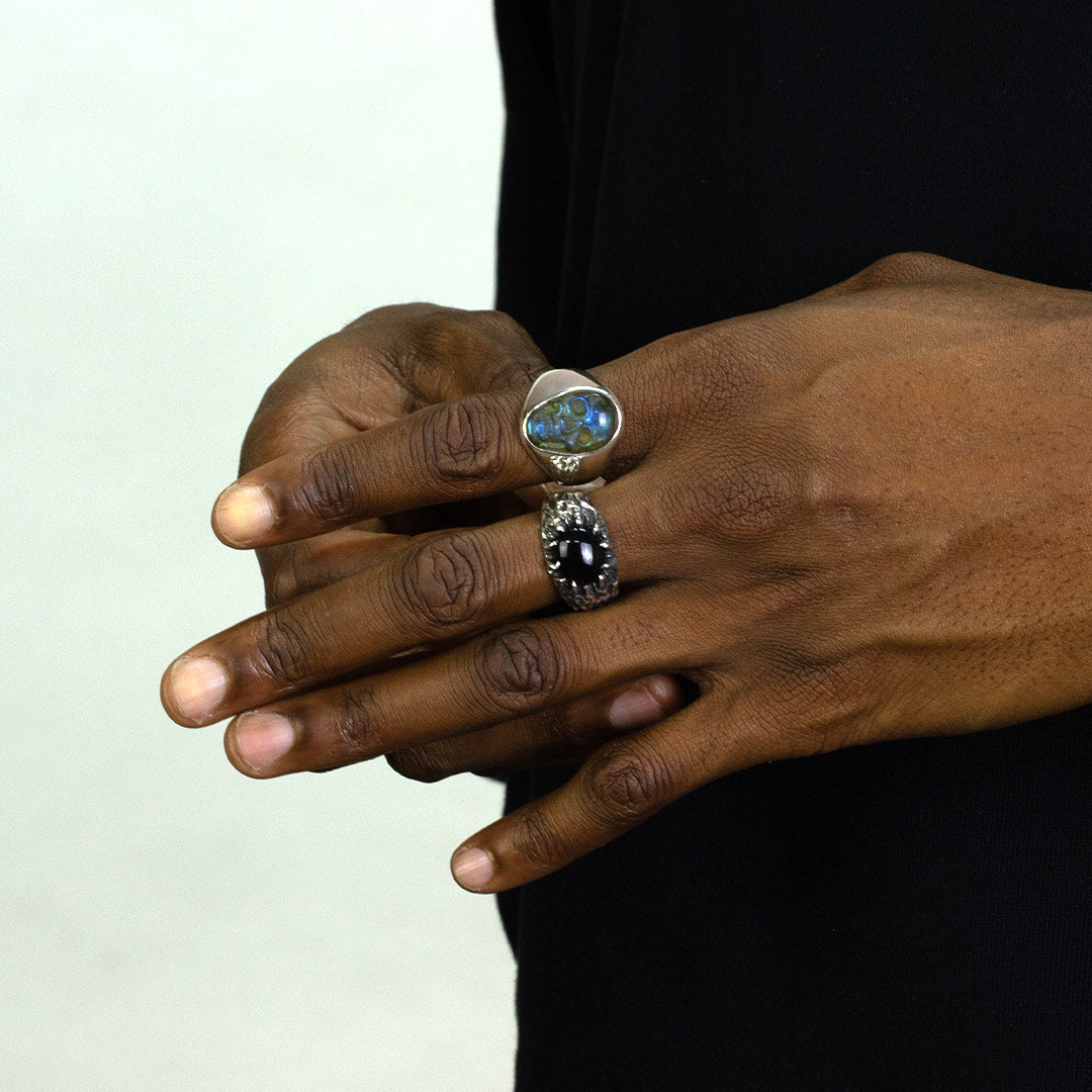 Close-up of a person's hands wearing a silver ring with a black gemstone and textured band on a plain background