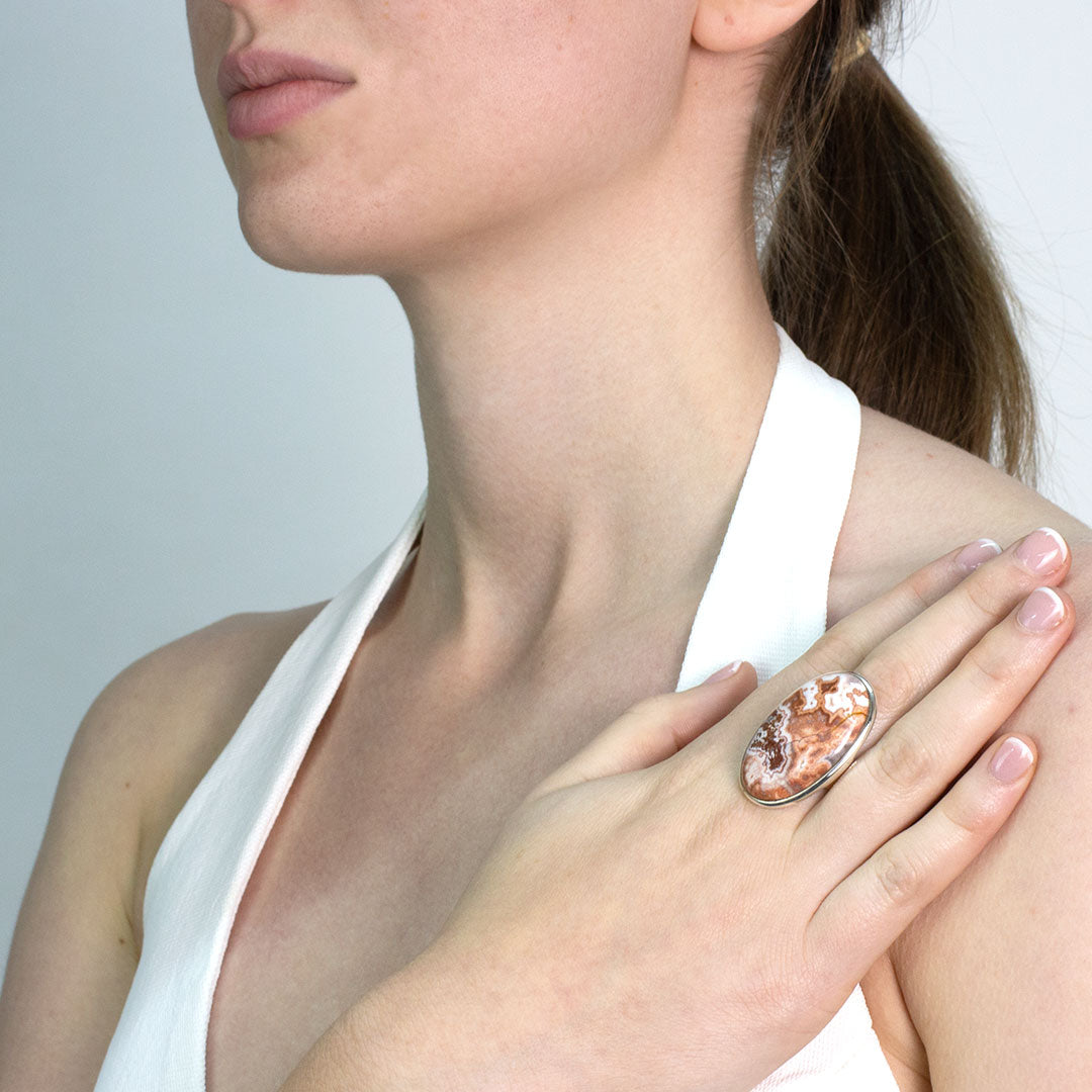 Woman wearing a silver ring with an oval stone with brown, orange and white patterns on a plain background