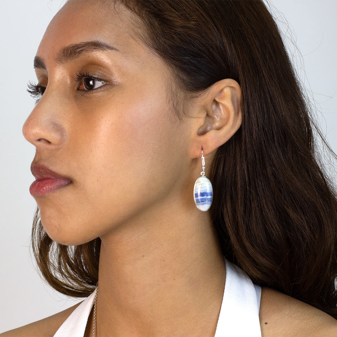 Woman wearing an oval-shaped silver earring with blue and white striped stone on a plain background
