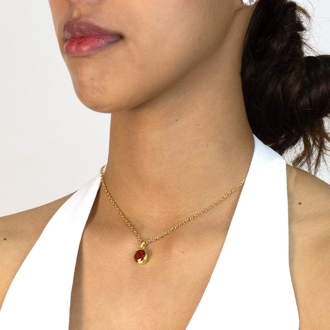 Woman wearing a gold necklace with an oval orange gemstone pendant on a plain background