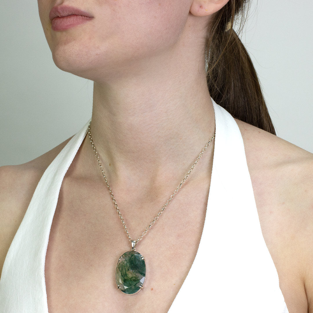 Woman wearing a silver necklace with translucent gemstone with green inclusions on a plain background