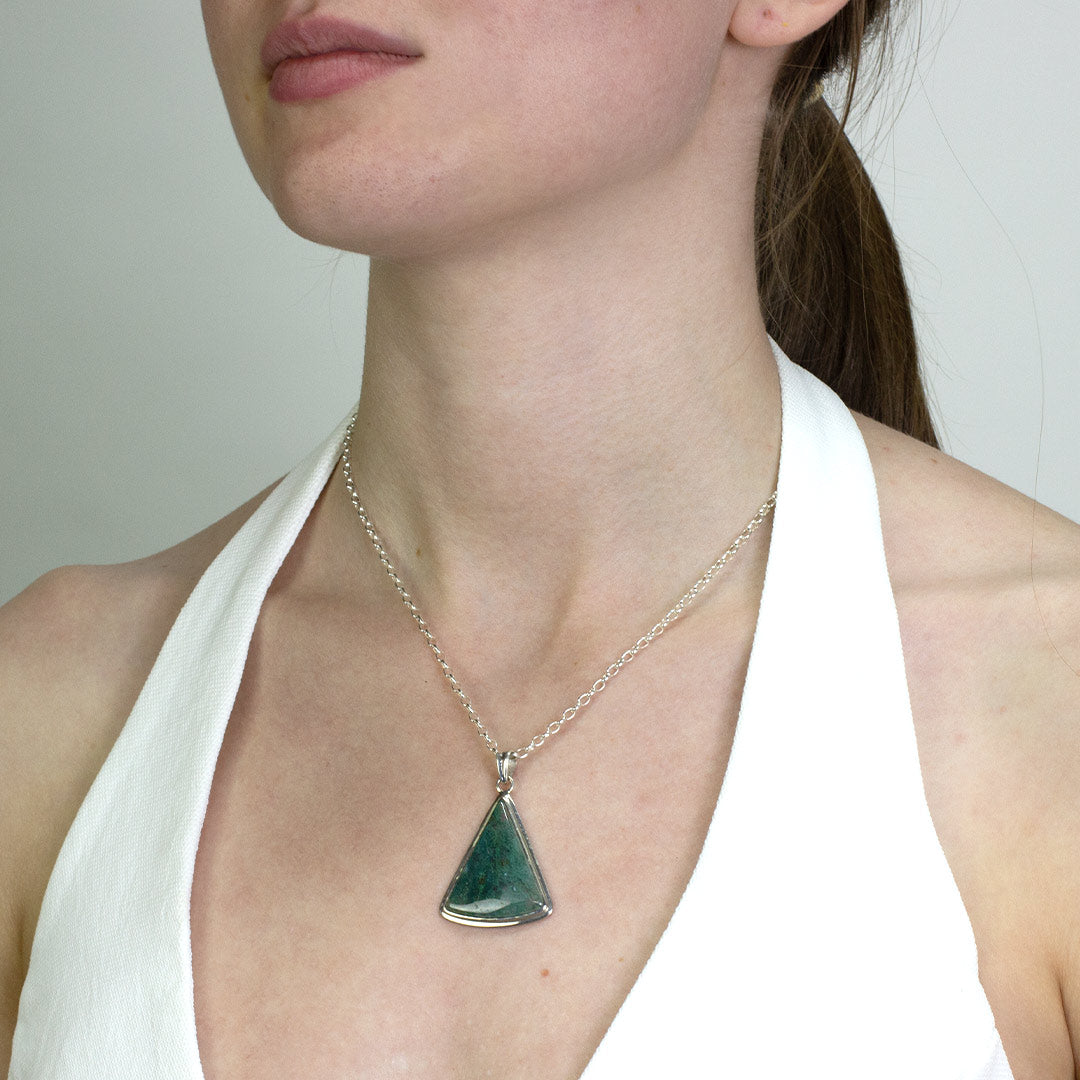 Person wearing a silver necklace with translucent gemstone with green inclusions on a plain background
