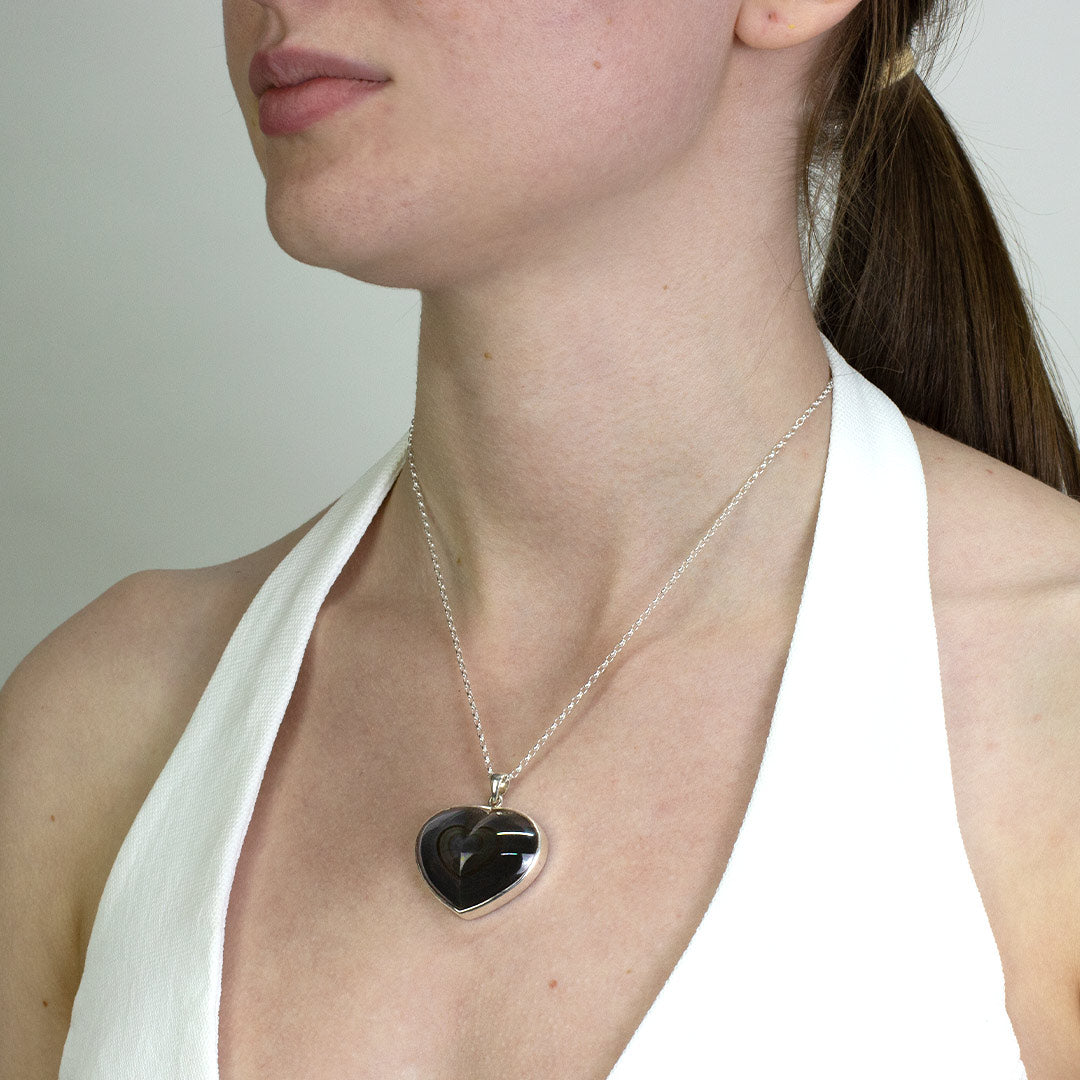Woman wearing a silver necklace with a black heart-shaped pendant on a plain background
